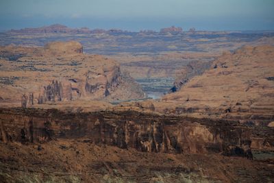 Telephoto view from a high rim across a maze of slickrock domes and fins to the Colorado River emerging through the Portal, with hazy sandstone towers on the far horizon.