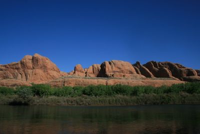 Red slickrock domes and fins lit by morning sun above a band of green cottonwoods, with the shaded Colorado River flat in the foreground.