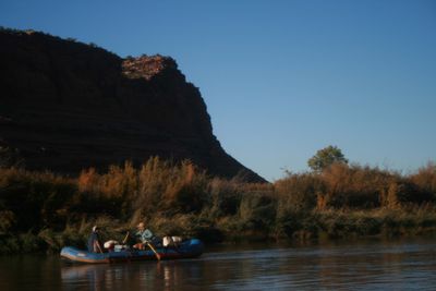 Two boaters in a loaded blue raft rowing close to a bank of fall-colored tamarisk and willows