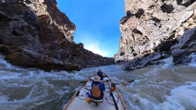 View from the bow of a dory as a rower in a straw hat and orange life vest runs whitewater between steep black rock walls in Westwater Canyon, with a second boater seated at the front hatch under a clear blue sky.
