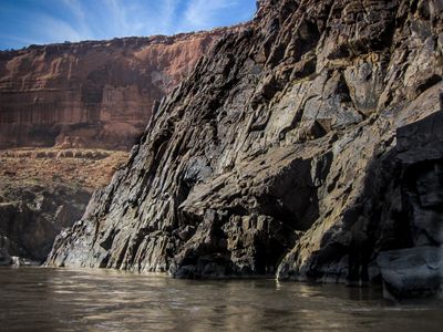 Fractured black metamorphic cliff dropping into brown water in Westwater Canyon above Skull Rapid, with red sandstone walls and caprock formations in the distance.