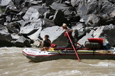 A rower in a straw hat and red life vest stands at the oars of a loaded white gear raft on muddy brown water in Westwater Canyon, with a woman seated on the bow tube and a jumble of grey boulders behind.