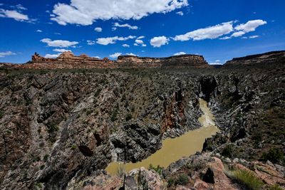 View from the rim into Westwater Canyon's inner gorge, where the muddy Colorado River bends between black rock walls below red sandstone buttes and a blue sky with scattered clouds.