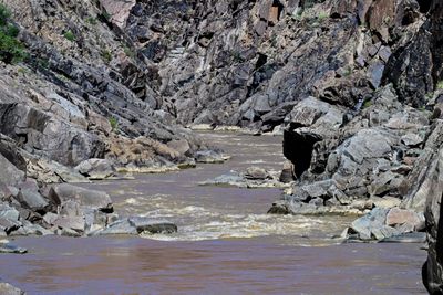 Muddy brown rapids of the Colorado River threading between fractured black boulders and steep dark gorge walls in Westwater Canyon.