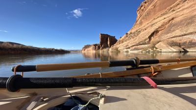 Wooden oars rest across the deck of a dory on calm reflective water in the open sandstone miles of the Westwater Canyon stretch, with a red sandstone tower and low brushy banks downstream under a deep blue sky.
