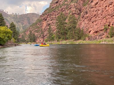 Paddlers in blue and yellow rafts float a wide green pool of the Green River's A Section as rain dimples the water beneath the red cliffs and ponderosa pines of Red Canyon.