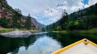 View over the bow of a white dory with a yellow gunwale gliding on glassy green water between red canyon walls and ponderosa pines under building cumulus clouds.
