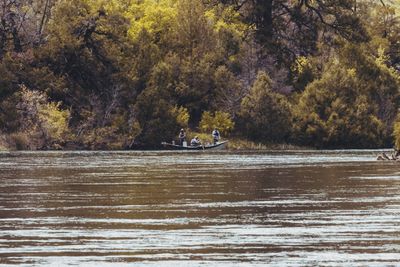 A drift dory tucked along the far brushy bank of the Green River, with two anglers standing to cast fly rods and a rower at the oars, seen across broad riffled current.