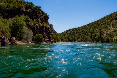 Sunlight sparkles across riffled turquoise current below a shaded red rock cliff and juniper-covered slopes under a cloudless sky on the Green River's A Section.