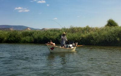 A person in a straw hat stands casting a fly rod from a wooden dory on the green Green River, a second person seated in the bow
