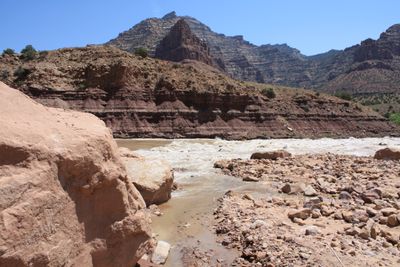 Silty whitewater breaks across the full width of the Green River at Cow Swim Rapid, viewed from a rocky debris fan with a large sandstone boulder in the left foreground and dark banded cliffs rising to a pyramid-shaped peak behind.