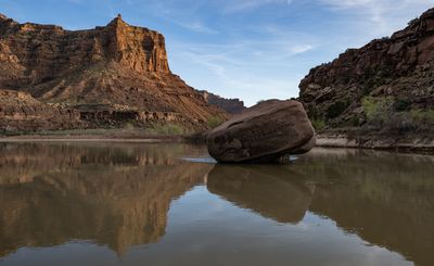 A massive rounded boulder rests at the calm edge of the silty Green River in Desolation Canyon, mirrored in the still water, with a warm-lit butte and layered canyon walls upstream under wispy clouds.