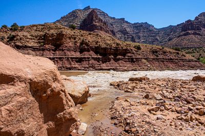 Whitewater churns across the silty Green River at Cow Swim Rapid, seen from a boulder-covered debris fan with a tall sandstone block in the left foreground and maroon banded slopes rising to a pyramidal peak beyond.