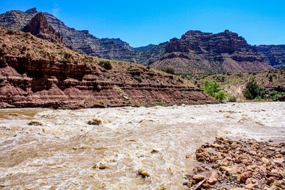 A long stretch of frothing brown whitewater fills the Green River at Cow Swim Rapid, with a cobble bar in the right foreground and stepped maroon cliffs and desert slopes rising behind under a deep blue sky.