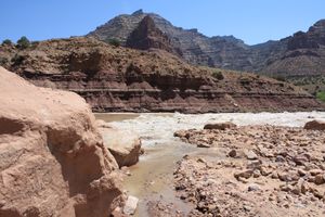 Rock Creek Rapid running silty and big at high water on the Green River in Desolation Canyon, seen from a boulder-strewn debris fan where a clear side stream trickles in, below banded red-brown slopes and a pointed layered peak under blue sky.