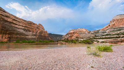 A pink gravel bar at Echo Park where the Green River bends beneath the striped wall of Steamboat Rock, with green cottonwoods and willows on the far bank and rounded sandstone domes under a bright cloud-filled sky.