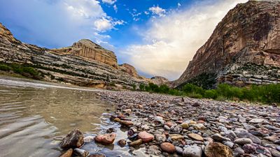 Wet, multicolored cobbles at the water's edge in Echo Park, with rain-dimpled water at left and the Green River curving between the tall face of Steamboat Rock and a rounded sandstone dome under dramatic clouds.