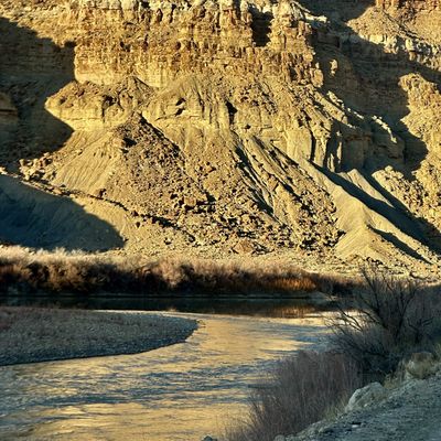 Evening sun rakes across a boulder-covered talus slope and layered cliff band in Gray Canyon, above a shadowed bend of the Green River reflecting gold along a gravel bar.