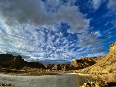 Streaked white clouds fan across a deep blue sky above the shadowed walls of Gray Canyon, with winter-bare cottonwoods on a gravel bar and the wide, silty Green River in the foreground.