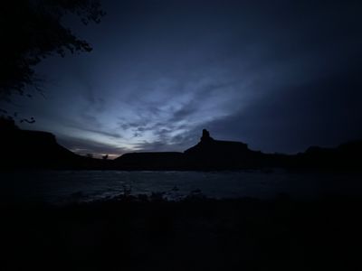 Gunnison Butte stands silhouetted against deep blue dusk clouds at the mouth of Gray Canyon, a single bright planet overhead and the Green River running dark in the foreground.