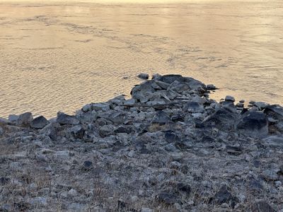 A point of gray boulders, dry grass, and driftwood juts into the rippled Green River at dusk in Gray Canyon, the silty water reflecting pale gold.