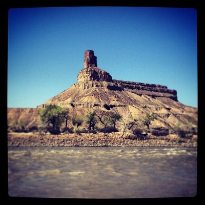 Gunnison Butte's chimney-topped mesa rises over green cottonwoods and the silty Green River in a heavily filtered, vignetted square snapshot.