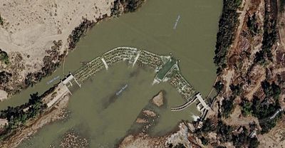 Satellite view of the low diversion weir angling across a bend of the silty Green River near the town of Green River, Utah, with whitewater spilling over the crest, exposed gravel below the dam, and Hastings Road following the tree-lined east bank.