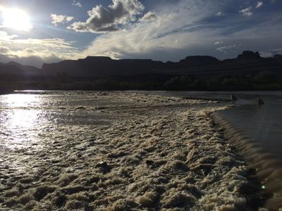 Silty whitewater churns over the Green River diversion weir in the town reach, backlit by a low sun breaking through clouds, with power lines crossing and dark bluffs silhouetted on the skyline.