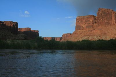 Late-afternoon sun on sandstone buttes and talus slopes above the Green River in Labyrinth Canyon, with reeds and willows along the far bank.