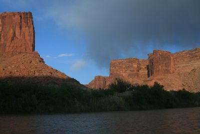 Sandstone tower and cliffs above the reed-lined bank of the Green River in Labyrinth Canyon, with dark storm haze moving in behind.