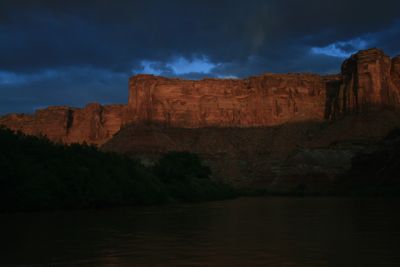 Last light glowing on a sandstone cliff band in Labyrinth Canyon while the Green River and its banks fall into storm-dark shadow.