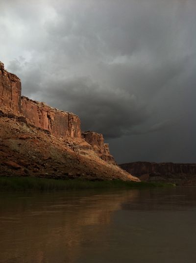 Sunlit red cliffs reflected in the calm surface of the Green River under a dark storm sky in Labyrinth Canyon.