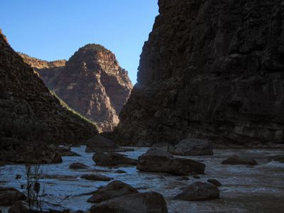 Silty water threads between large boulders in a deeply shaded stretch of Lodore Canyon, with a sunlit red peak rising beyond under a clear blue sky.