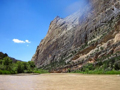 The sheer, streaked face of Steamboat Rock rises straight from the muddy Green River near Echo Park, with a low sandbar and green banks at its base.