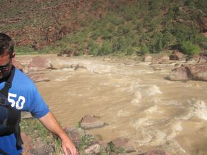 A rafter in a blue shirt and sunglasses picks his way along the rocky bank while scouting Hell's Half Mile rapid, its silty brown water breaking over a wide boulder garden in Lodore Canyon.