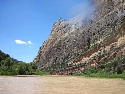 Wide view of Steamboat Rock's sheer sandstone face towering over the muddy Green River near Echo Park, with a sandbar and brushy banks in the foreground.