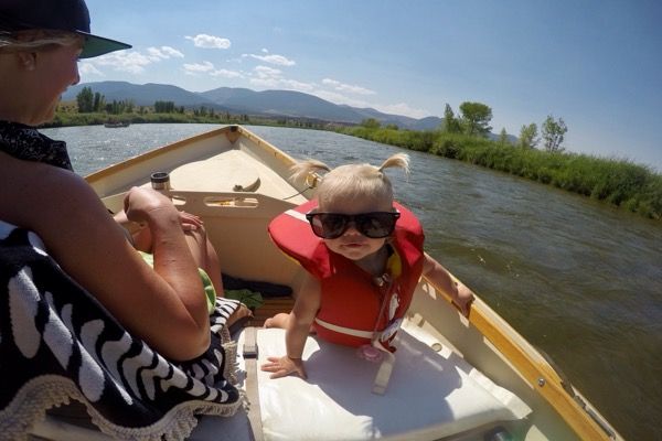 A toddler in a red life jacket and oversized sunglasses stands at the front seat of a small dory while an adult looks
