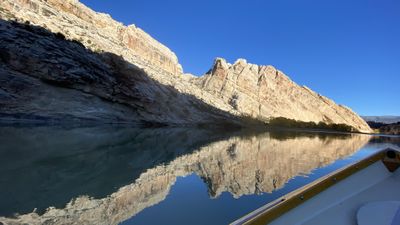 Mirror-calm Green River reflecting the tilted, striped cliffs of Split Mountain Canyon under a deep blue sky, with the wooden gunwale of a dory at the lower right.