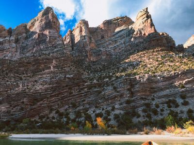 Tilted rock towers of Split Mountain rise above a white sand beach with rust-orange autumn shrubs at the river's edge, with the tip of a boat's bow entering the frame below.
