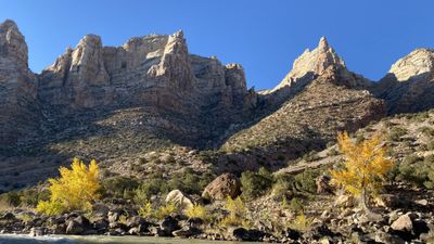 Sun lights the sculpted white crags of Split Mountain above a shadowed slope, with golden cottonwoods and boulders along the riverbank below.