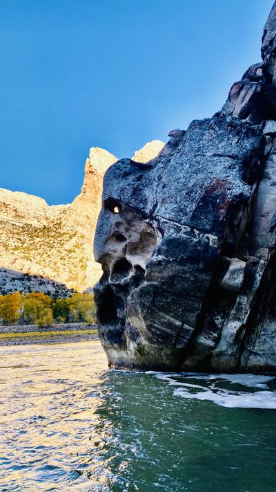 A dark, water-pocketed boulder face rises directly from the swirling green water of the Green River, with a sunlit crag of Split Mountain glowing behind it in fall.