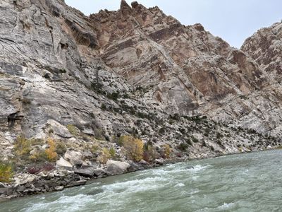 A towering fractured sandstone wall streaked with dark stains rises above a juniper-dotted talus slope and the fast green water of the Green River in fall.