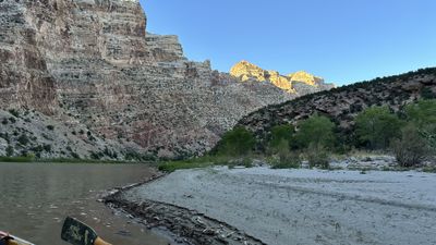 Last light catches the high rim of Split Mountain Canyon above a shadowed wall, with a sandy bank, green trees, and an oar blade resting over the muddy river edge in the foreground.