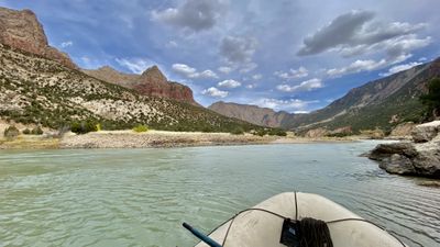 The bow of a tan raft with a coiled bow line drifts a calm, green stretch of the Green River toward the gates of Split Mountain under scattered clouds.