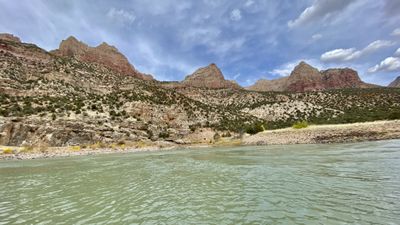Red-striped crags and pinnacles rise above a juniper-covered slope and yellow rabbitbrush at the edge of the Green River, under a blue sky with wisps of cirrus.