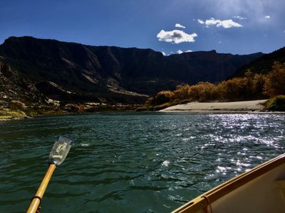 Sunlight sparkles on the dark green Green River as a dory's oar reaches toward a white sand beach backed by orange autumn cottonwoods and shadowed canyon walls.