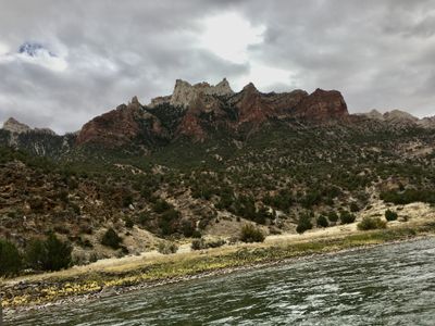 The jagged red-and-white crest of Split Mountain stands under a heavy overcast sky, above juniper slopes and a bank of yellow rabbitbrush at the dark, choppy river.