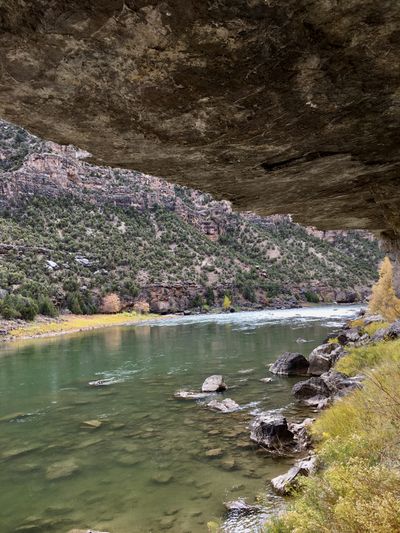View from beneath a massive overhanging rock roof at the river's edge, looking upstream at a riffled bend of the Green River with yellow brush and juniper slopes beyond.