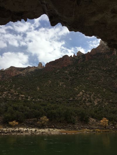 From the shade of an overhanging cliff, red-banded slopes dotted with junipers rise above a cobble shore and the green water of the Green River under a partly cloudy sky.