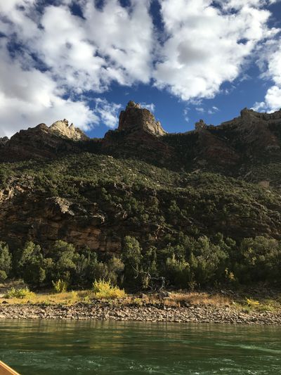 A pinnacled red-and-white ridge rises above dense juniper slopes and a cobble bank along the green water of the Green River.